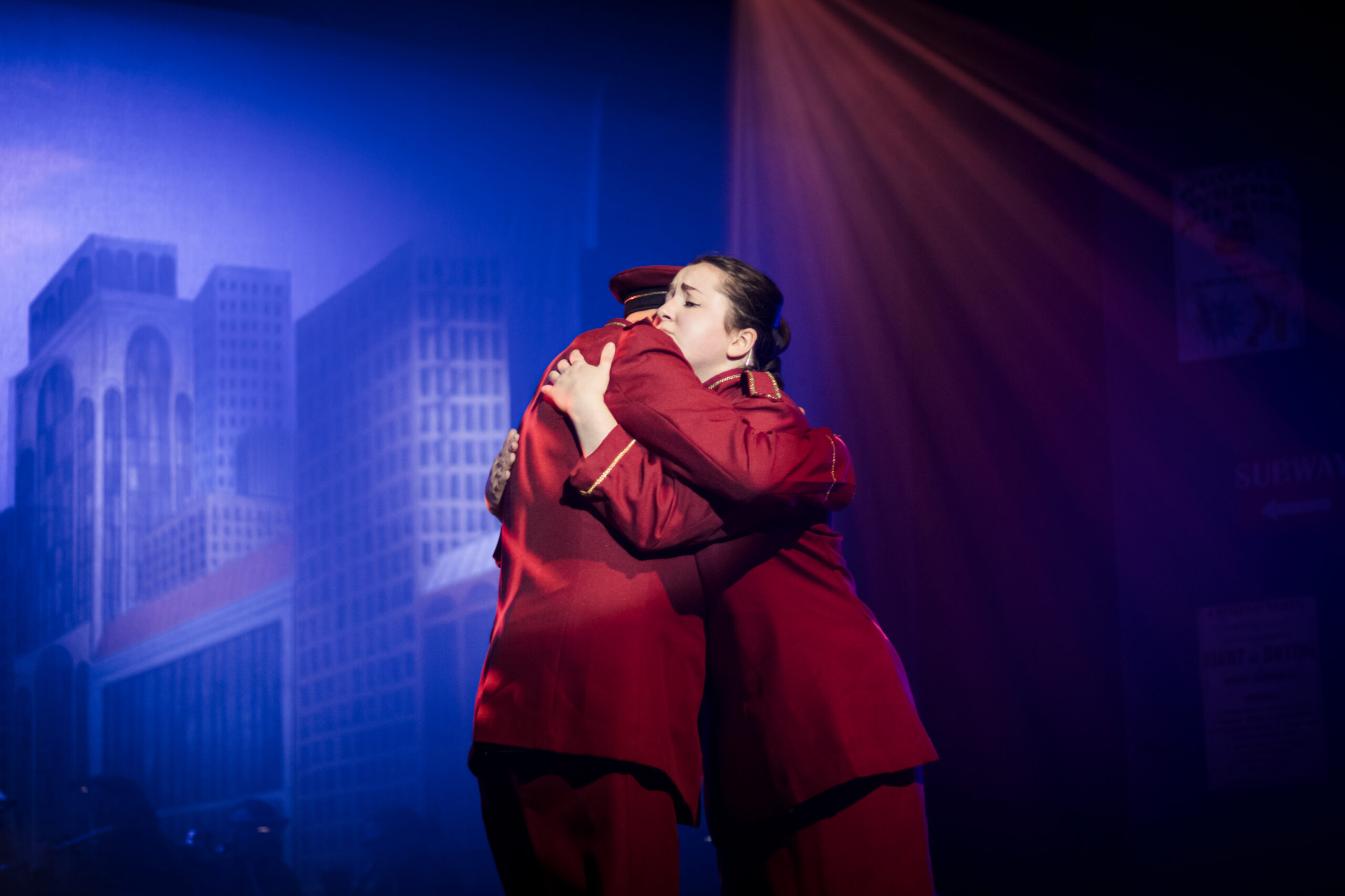 Emotional stage performance: Actors embrace against a cityscape backdrop with red lighting.