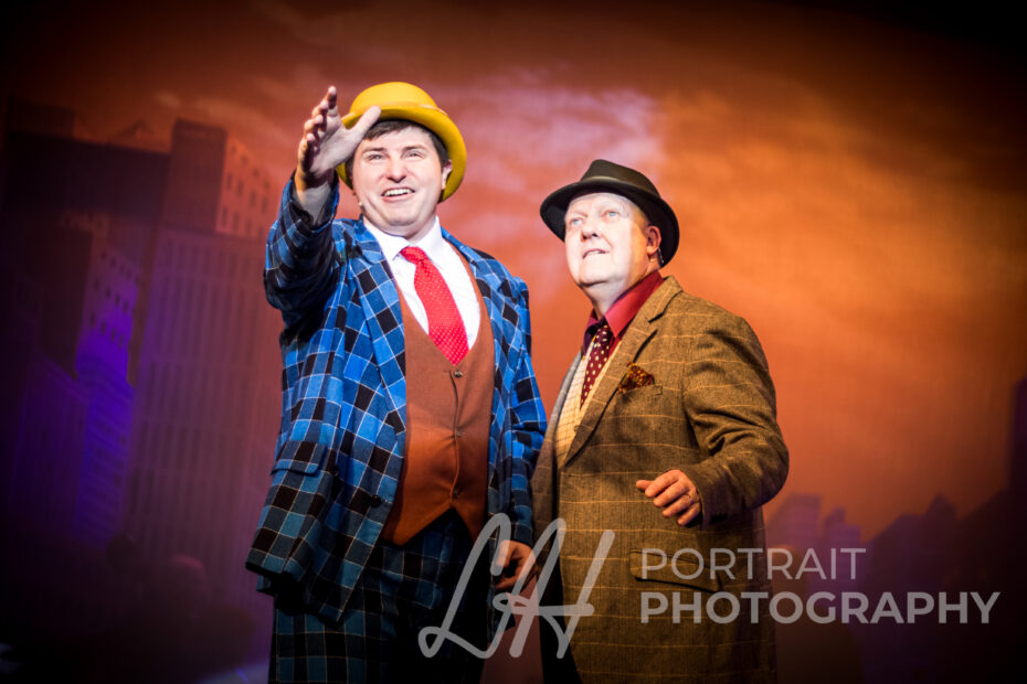 Stage photography: Two actors in costume, one in a bowler hat, performing on stage with city backdrop. Performance art.