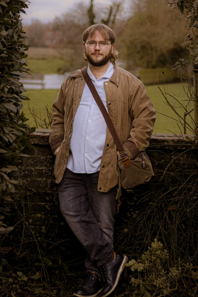 Professional photographer portrait. Man with glasses, jacket, and bag poses outdoors. Photography services available.