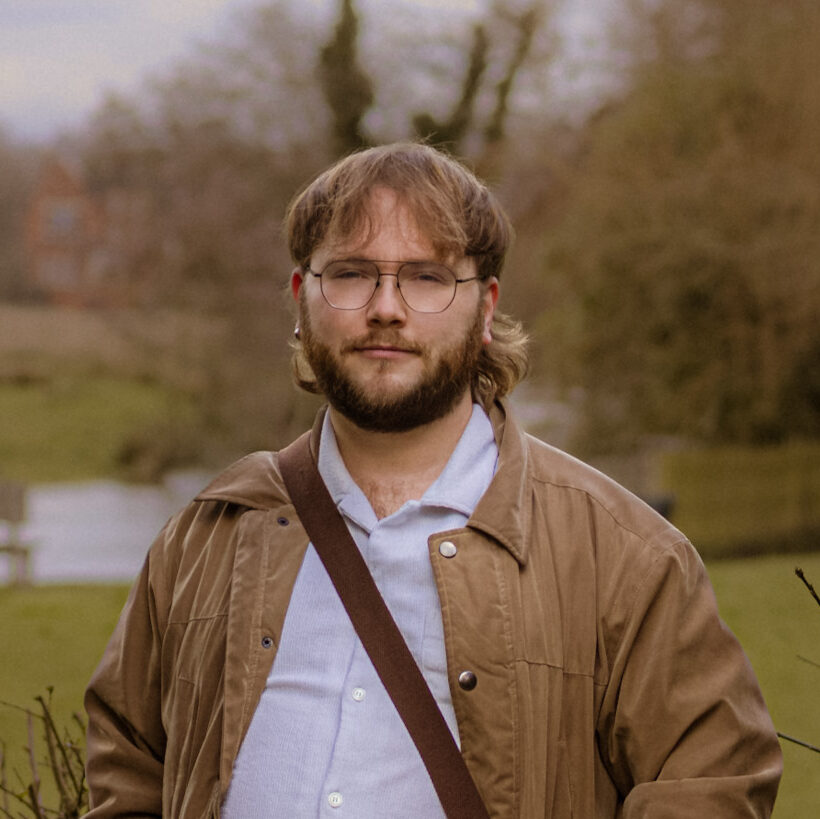 Professional photographer portrait. Man with glasses, jacket, and bag poses outdoors. Photography services available.